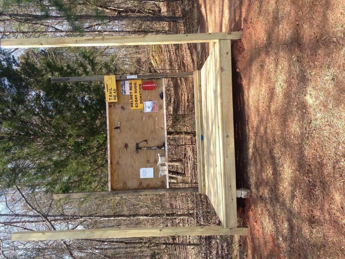 Wooden structure in a forested area, featuring a sign labeled "Trail Head." The structure includes a tabletop with various papers and a small faucet, with trees and a natural ground surface visible in the background. Rocky River Trail mountain bike trail.