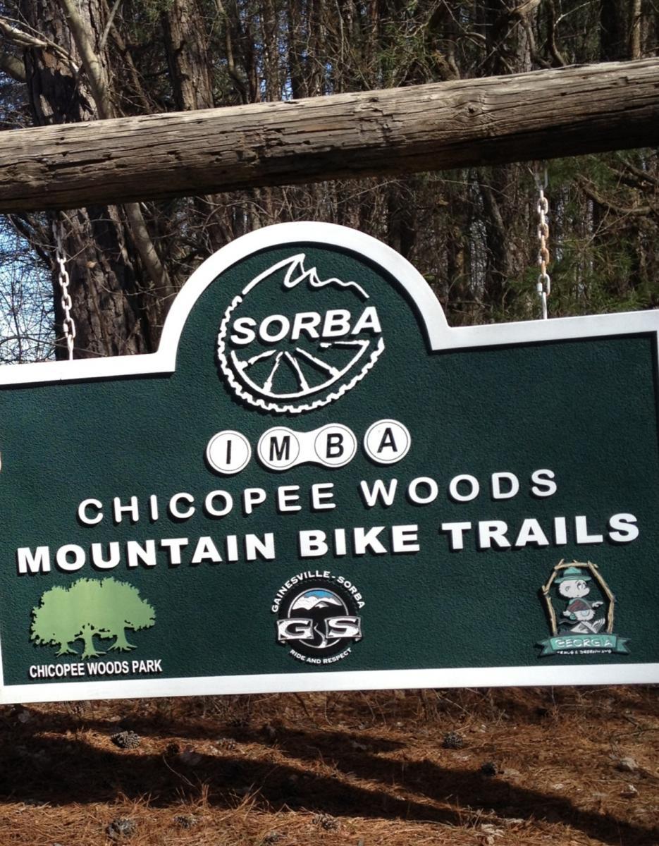 Sign indicating the entrance to the Chicopee Woods Mountain Bike Trails, featuring the logos of SORBA (Southern Off-Road Bicycle Association) and IMBA (International Mountain Bicycling Association), along with the name "Chicopee Woods Park" and associated local organizations. The sign is mounted on a wooden beam and surrounded by trees.