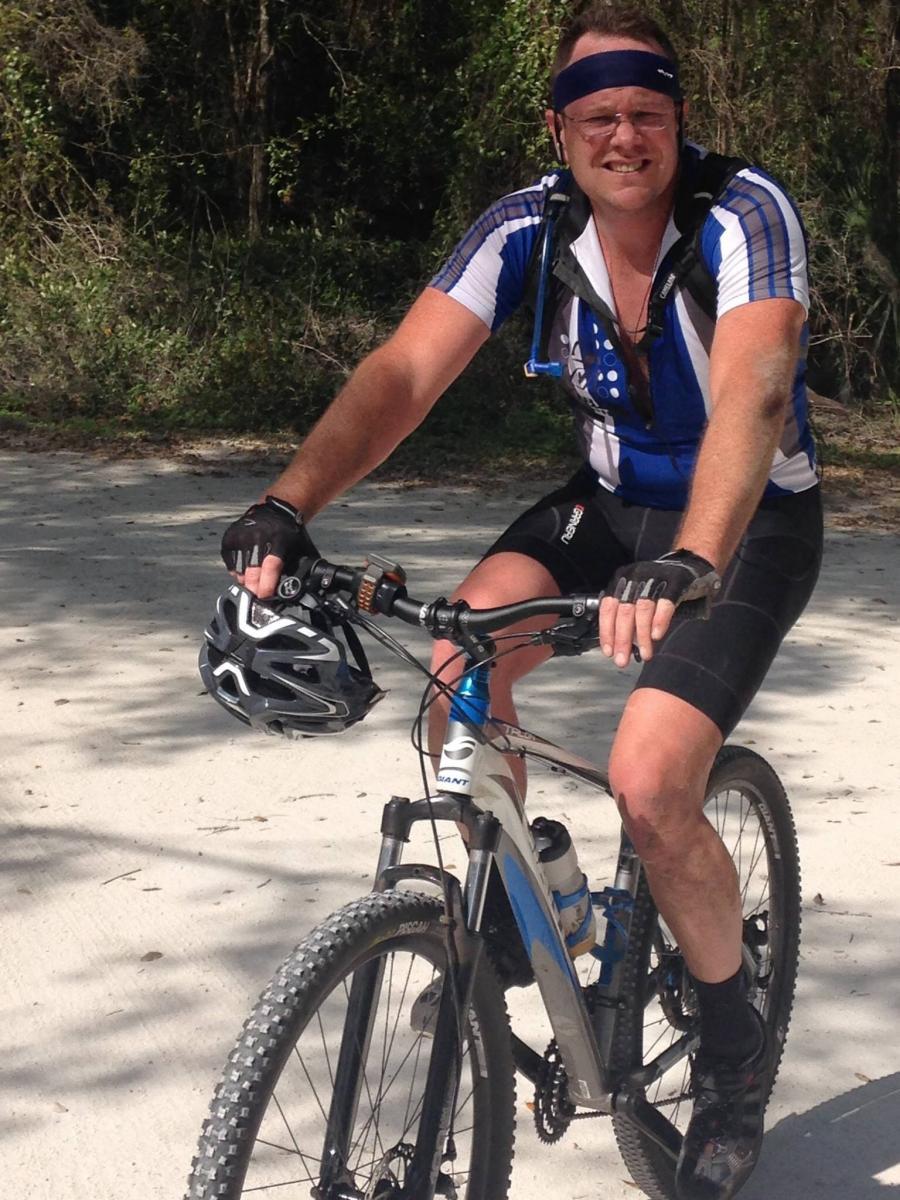 Giant Talon 29er 2: A cyclist dressed in a blue and white jersey, wearing a headband and sunglasses, smiles while riding a mountain bike on a gravel trail surrounded by greenery.