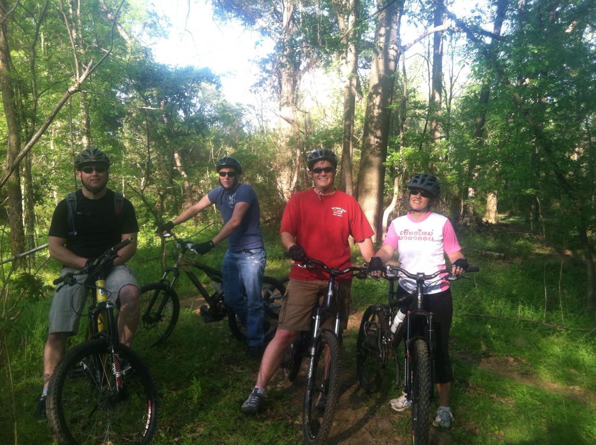 A group of four people, all wearing helmets and biking gear, are standing with their mountain bikes on a wooded trail. The sun filters through the green trees, creating a bright and natural setting. Two men and two women smile at the camera, enjoying their time outdoors. One man is wearing a red shirt, while another is in a blue shirt. The women are in a pink and black outfit. The scene captures a moment of camaraderie during an outdoor biking adventure. Spadra Creek Nature Trail mountain bike trail.