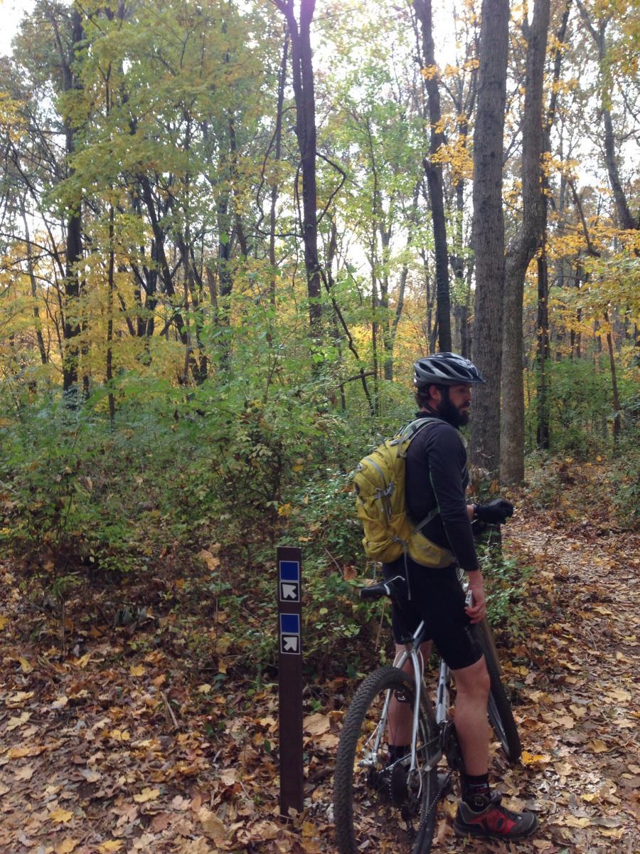 A person in cycling gear stands next to a mountain bike on a forest trail, surrounded by colorful autumn foliage and trees. A trail marker with blue signs indicating direction is visible nearby. Percy Warner Mountain Bike Trails mountain bike trail.