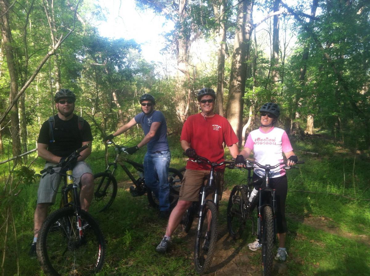 Four people in helmets are standing next to their mountain bikes on a wooded trail. They are smiling and enjoying a sunny day outdoors among greenery. Trees surround them, creating a natural setting for their biking adventure. Spadra Creek Nature Trail mountain bike trail.