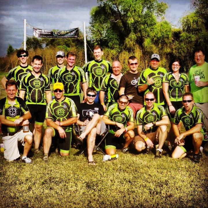 A group photo of 18 individuals wearing matching bright green and black cycling jerseys, posing together outdoors. They are smiling and standing in front of a tree line with a banner in the background. The setting appears to be a cycling or outdoor event, with some participants holding drinks.
