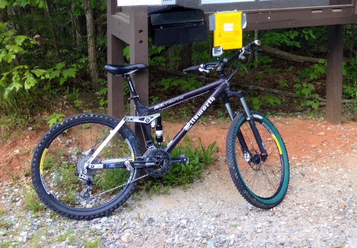 Ellsworth Truth: A mountain bike resting on gravel near a trailhead sign, surrounded by greenery. The bike features a black and silver frame with a green accent on the wheels. A nearby information board displays various notices and maps.