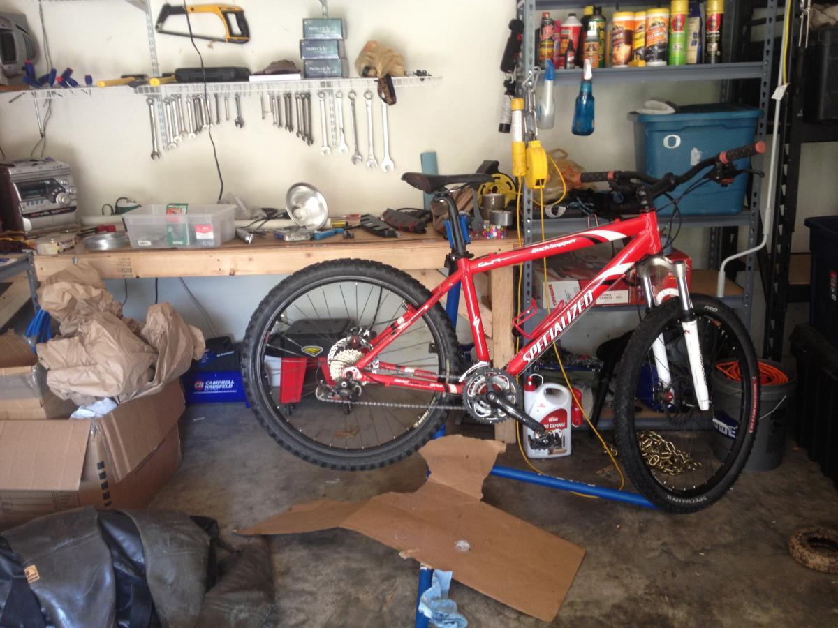 Specialized Rockhopper: A cluttered garage workspace featuring a red mountain bike leaning against a wooden workbench. The bench is covered with various tools, including wrenches and containers, while shelves above hold additional tools and supplies. There are several cardboard boxes and a black bag on the floor, creating a busy and utilitarian atmosphere.