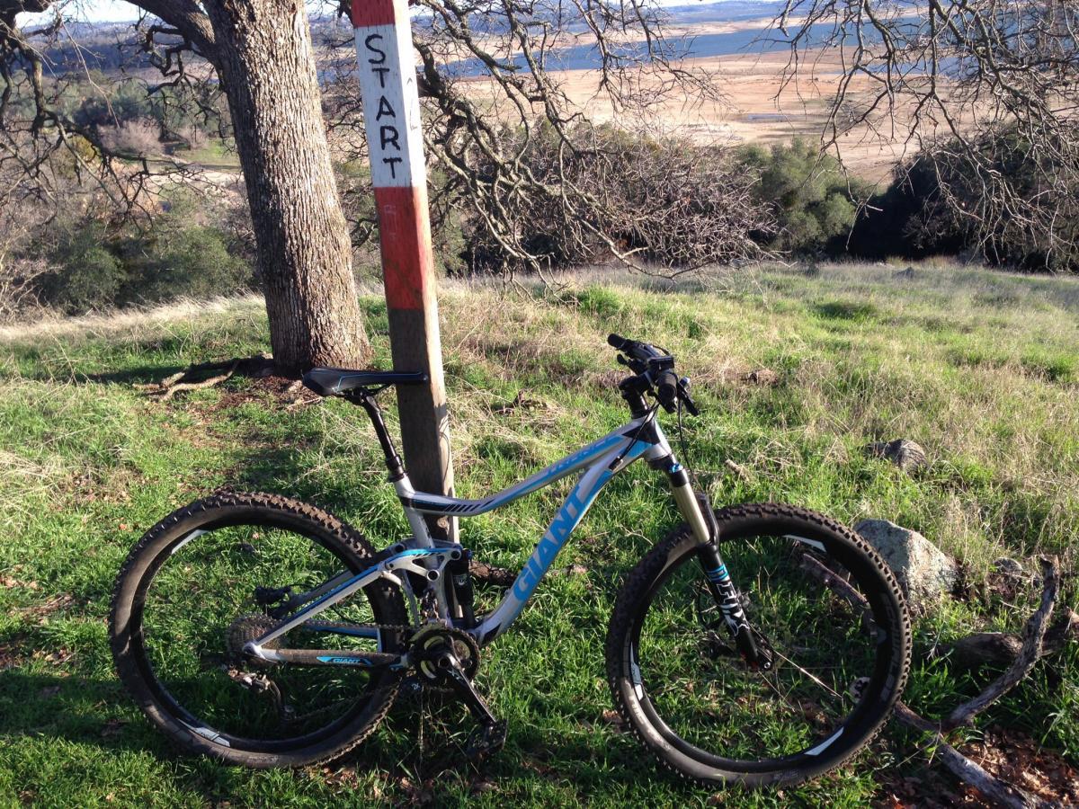 Giant Trance 27.5 1: A mountain bike parked on green grass next to a wooden post marked "START," with a scenic view of hills and trees in the background.