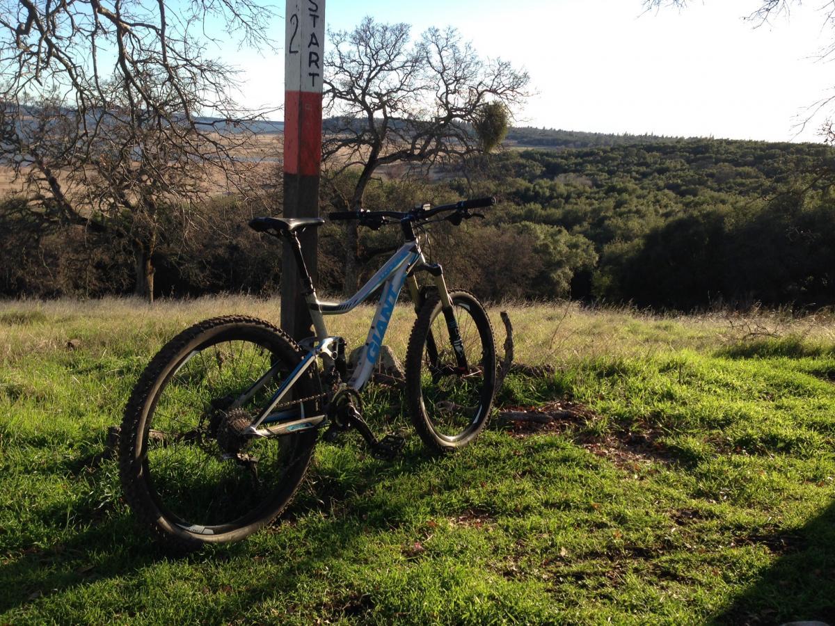 Giant Trance 27.5 1: A mountain bike resting on a grassy hillside, next to a trail marker indicating the start of a biking path. In the background, a view of rolling hills and trees under a clear sky. The bike is partially in shadow, showcasing its design and features.