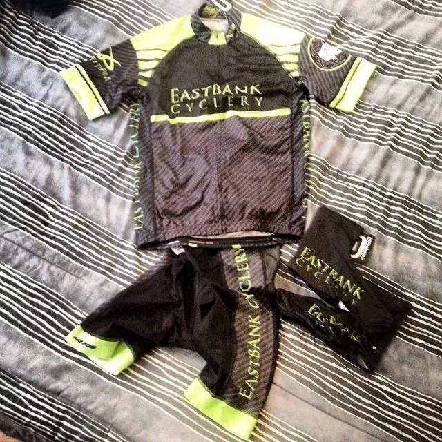 A cycling outfit laid out on a striped bedspread, consisting of a black jersey with green accents featuring "Eastbank Cyclery" branding, matching black shorts, and a coordinating headband.