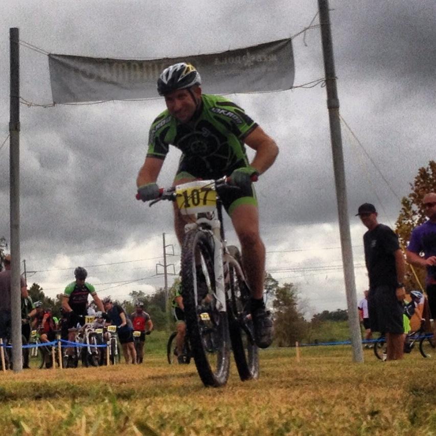 A mountain biker in a green and black racing outfit is riding toward the camera, crossing under a banner. The cyclist, wearing a helmet and number 107 on their bike, has a focused expression. In the background, other cyclists and spectators are visible, with overcast skies and grassy terrain.
