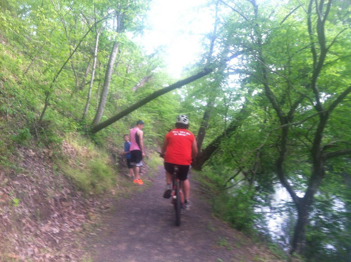 A scenic trail surrounded by lush greenery, with a cyclist in a red shirt riding on a path beside a body of water. Two individuals are seen walking on the trail, one in a pink tank top and the other in a blue shirt. Trees with leafy branches arch over the path, creating a serene outdoor atmosphere. Spadra Creek Nature Trail mountain bike trail.