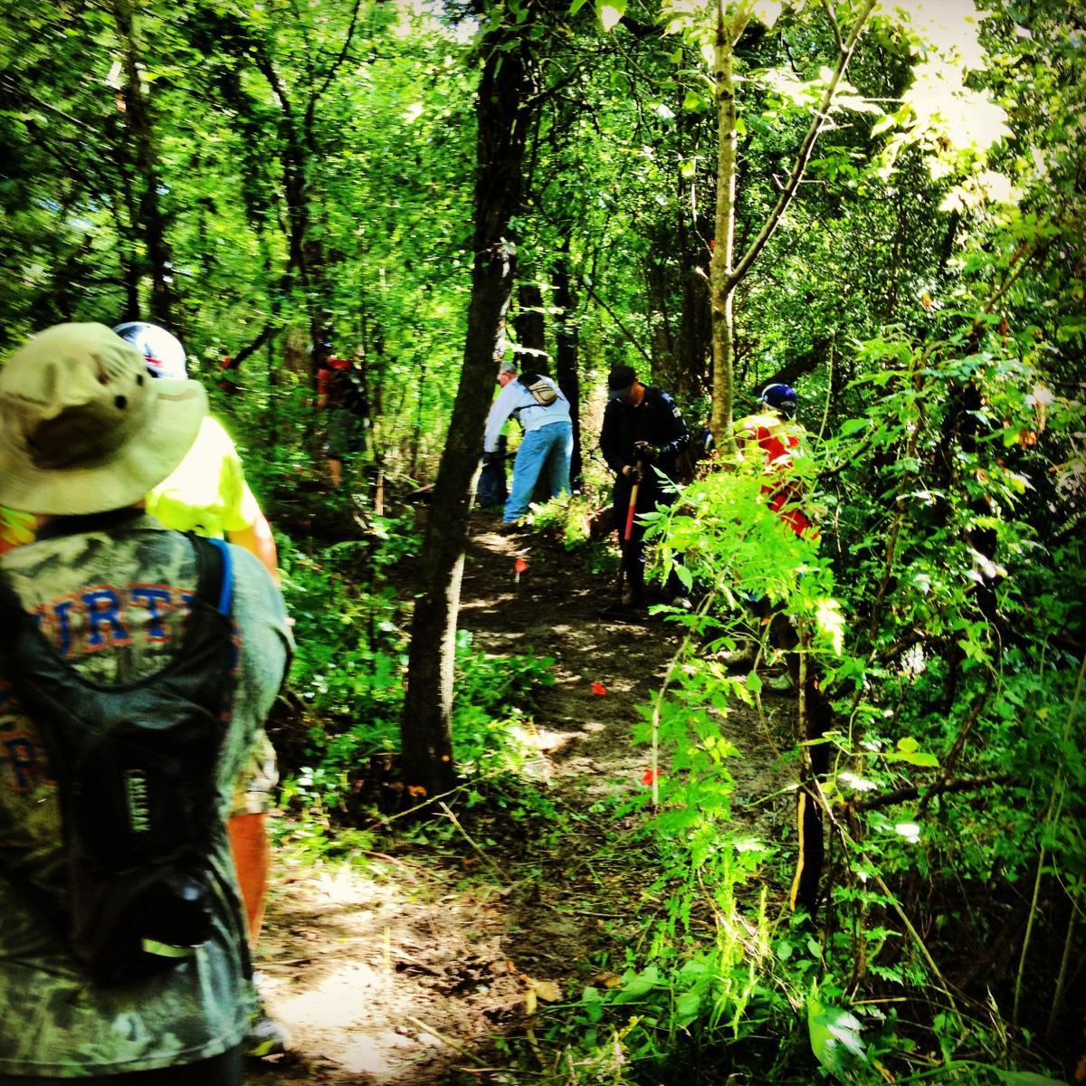 A group of people working on a wooded trail, surrounded by lush greenery. Some individuals are clearing the path, while others are marking it with flags. The scene captures teamwork in a natural setting. Bonnet Carre Spillway Trail mountain bike trail.