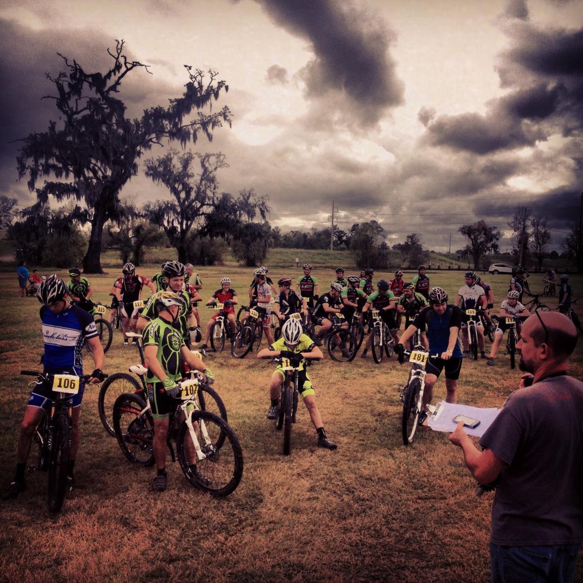 A group of mountain bikers gathered on a grassy field, preparing for a race. The participants, wearing various brightly colored jerseys and helmets, stand beside their bikes while listening to a man with a clipboard who is giving instructions. In the background, a dramatic sky with dark clouds looms overhead, adding to the atmosphere of the event. Bonnet Carre Spillway Trail mountain bike trail.