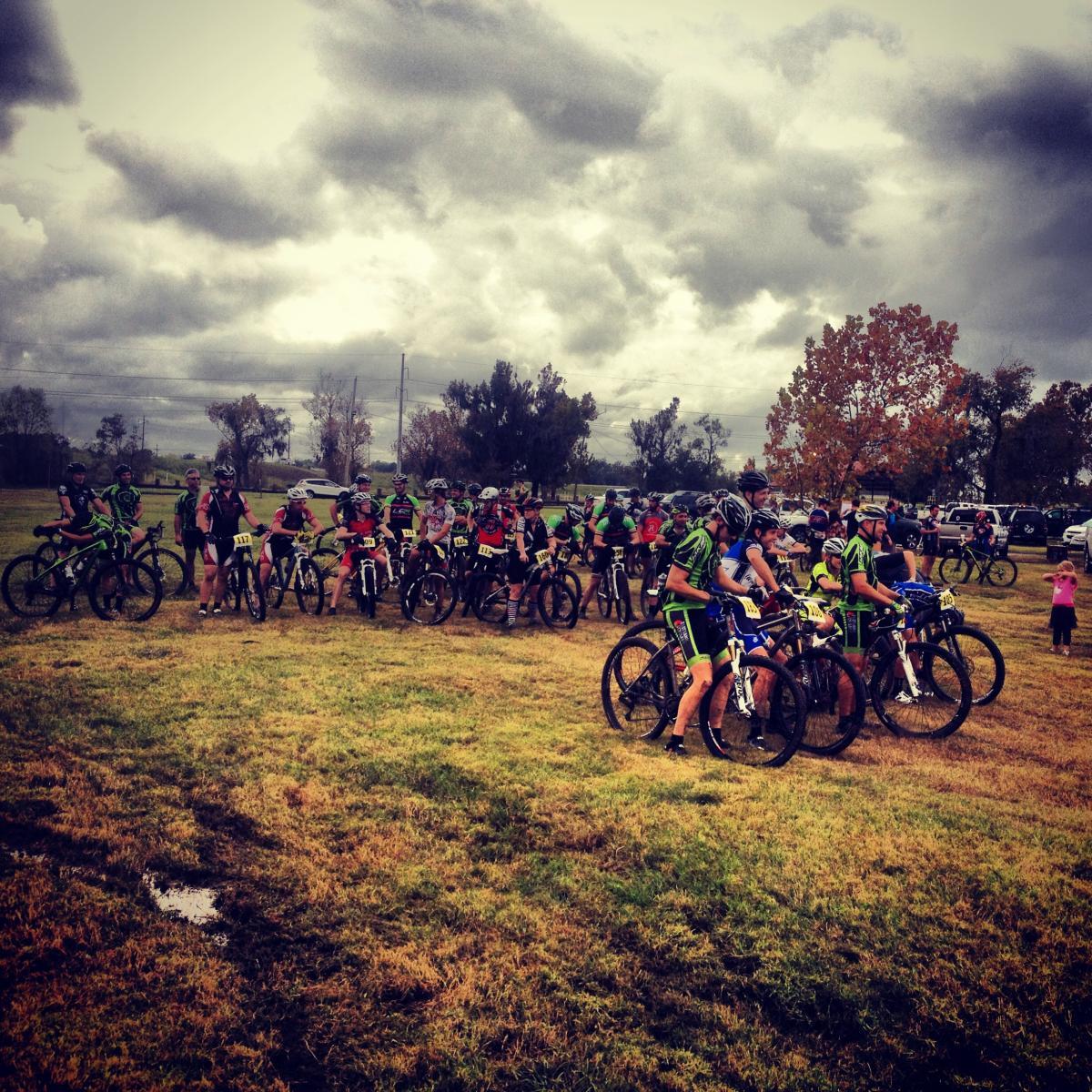 A group of cyclists in green and black jerseys gathers on a grassy field, ready for a race. The cloudy sky looms overhead, adding a dramatic backdrop to the scene. Some cyclists are seated on their bikes while others stand nearby, all preparing for the event. In the background, a few spectators and parked vehicles can be seen. Bonnet Carre Spillway Trail mountain bike trail.