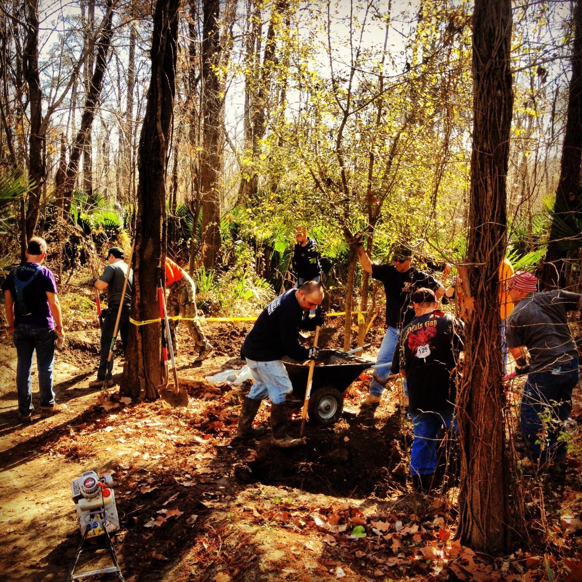 A group of people engaged in outdoor work in a wooded area, digging and preparing the ground. Some individuals are using shovels and wheelbarrows, while others are standing nearby. The setting features trees with bare branches and some greenery in the background. Sunlight filters through the trees, creating a bright atmosphere. There are tools and equipment scattered around, indicating a collaborative effort for a project in nature. Bonnet Carre Spillway Trail mountain bike trail.