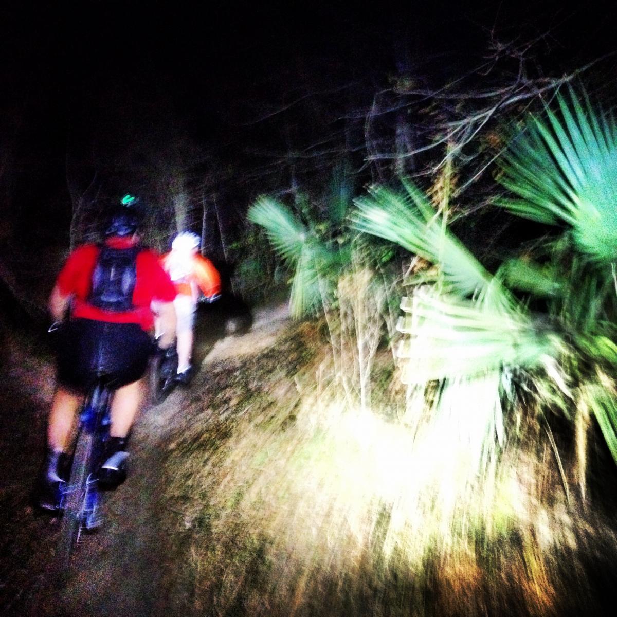 Two mountain bikers ride along a narrow, dimly lit trail at night, illuminated by their bike lights. The path is surrounded by dense vegetation, including palm leaves. The scene conveys a sense of adventure in a natural setting. Bonnet Carre Spillway Trail mountain bike trail.