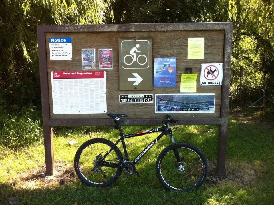 A mountain bike leaning against a wooden informational sign displaying rules and regulations for the Nomambo Bike Trail. The sign includes various notices, a map, and symbols indicating bike traffic, while a "No Horses" sign is also visible. Lush green grass and trees are in the background. Bonnet Carre Spillway Trail mountain bike trail.