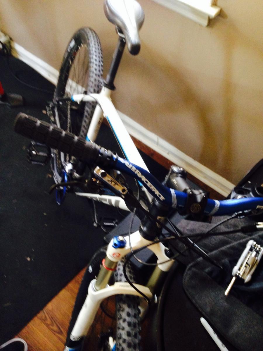 Trek X-Caliber: A close-up view of a mountain bike positioned on a dark carpeted floor, showcasing the handlebars, seat, and front suspension. The bike features a white frame with blue accents and has a dirty tire, indicating recent use. In the background, there are hints of other equipment and a light-colored wall.