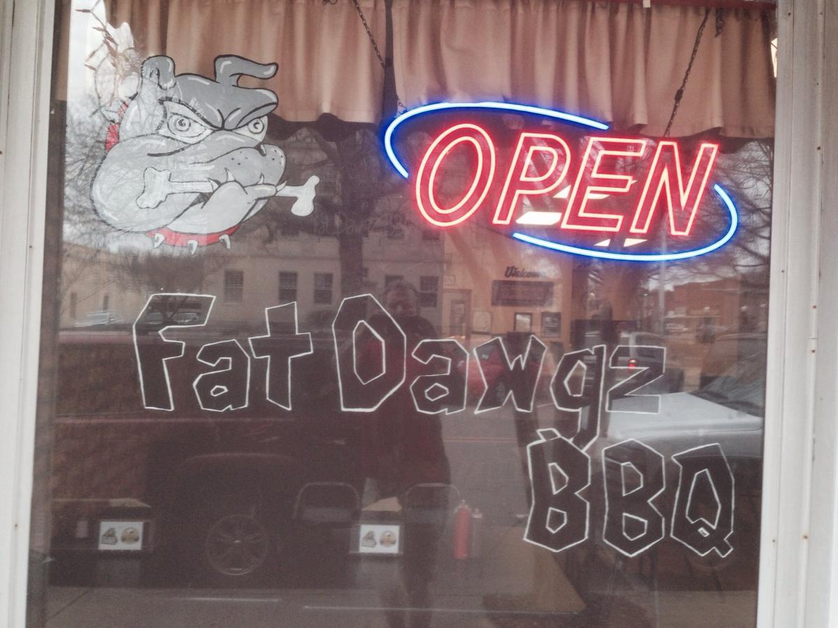 A storefront window displaying a vibrant neon "OPEN" sign, with a painted cartoon bulldog holding a bone and the text "Fat Dawgz BBQ" in bold, white lettering. The background features a light brown curtain. Spadra Creek Nature Trail mountain bike trail.