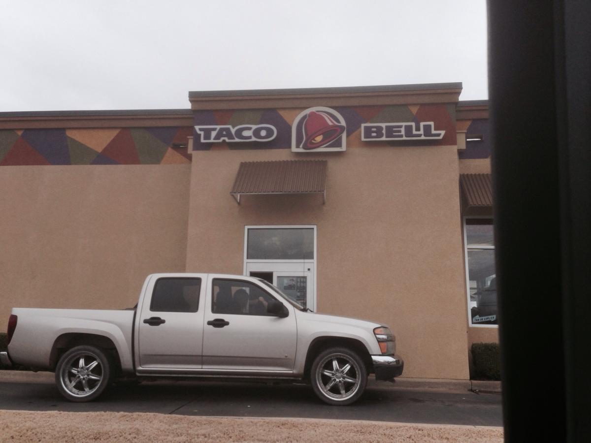 A white pickup truck parked outside a Taco Bell restaurant with a colorful geometric design above the sign, featuring the words "Taco Bell." The restaurant has a light brown exterior and a window displaying the service area. The sky is overcast. Spadra Creek Nature Trail mountain bike trail.