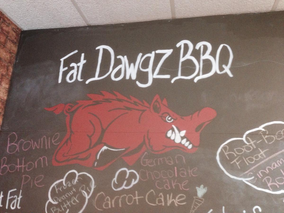 Wall mural featuring the text "Fat Dawgz BBQ" in stylized white lettering, accompanied by a vibrant red hog illustration. Below the text, various menu items are listed in colorful chalk, including "Brownie Bottom Pie," "Frozen Peanut Butter Pie," "Carrot Cake," "German Chocolate Cake," and "Root Beer Float." The background is a dark, chalkboard-like surface with a rustic texture. Spadra Creek Nature Trail mountain bike trail.