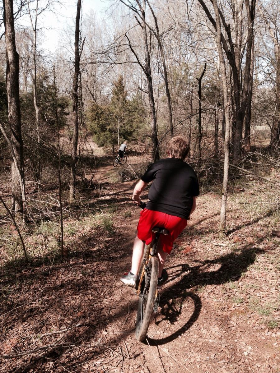 A young person riding a bicycle on a forest trail, with trees and underbrush surrounding the path. Another cyclist is visible in the background, navigating the same trail. The scene captures the tranquility of outdoor biking in a natural setting. Spadra Creek Nature Trail mountain bike trail.