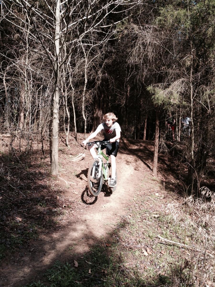 A young boy rides a green mountain bike along a dirt trail in a forested area, with trees surrounding him and dry leaves scattered on the ground. He appears to be caught in mid-action, enjoying a moment of biking through nature. Spadra Creek Nature Trail mountain bike trail.