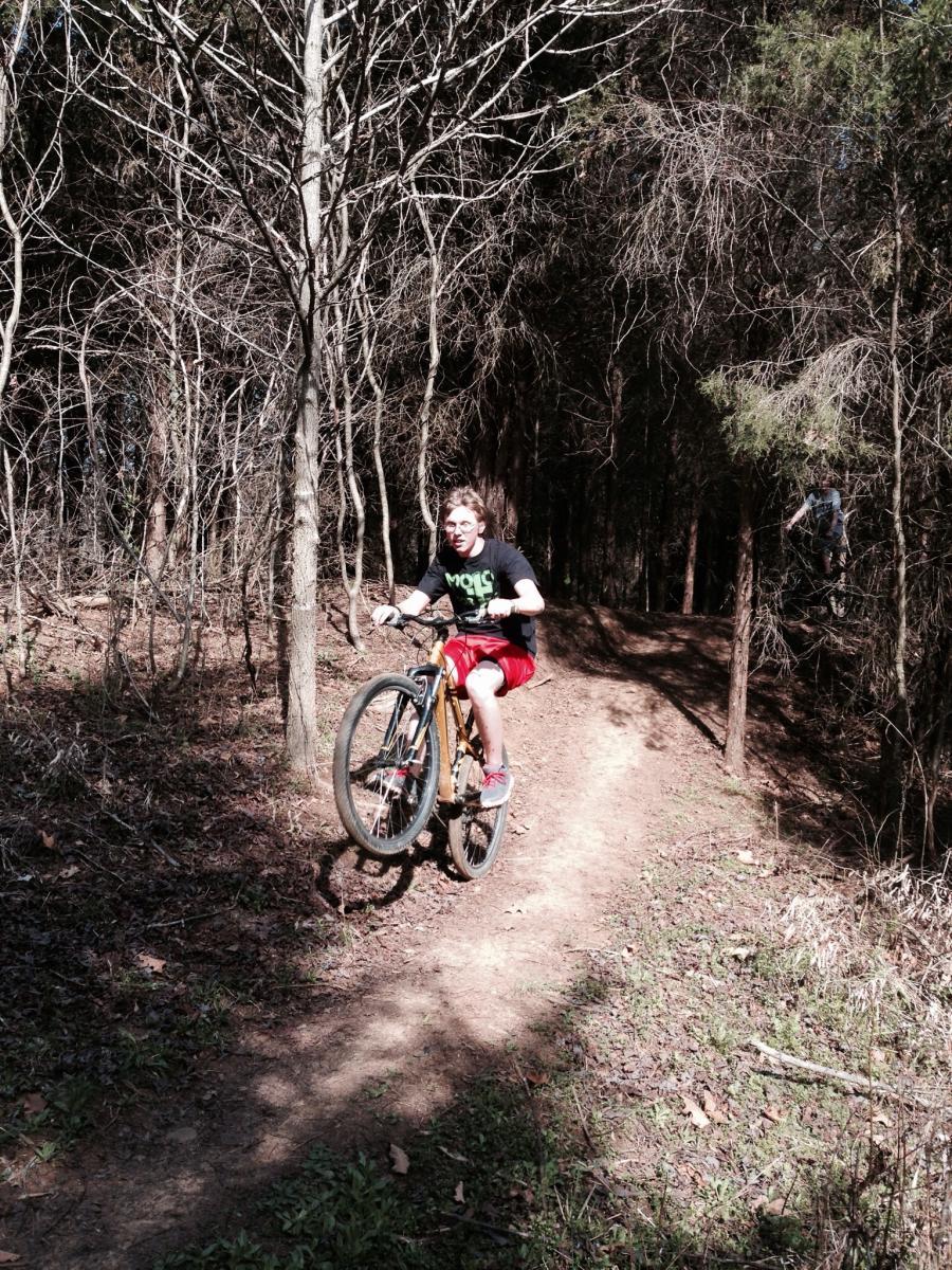 A child riding a bicycle on a dirt path in a wooded area, performing a wheelie. The scene is set among bare trees with the ground covered in leaves and grass, capturing the excitement of outdoor biking. Spadra Creek Nature Trail mountain bike trail.