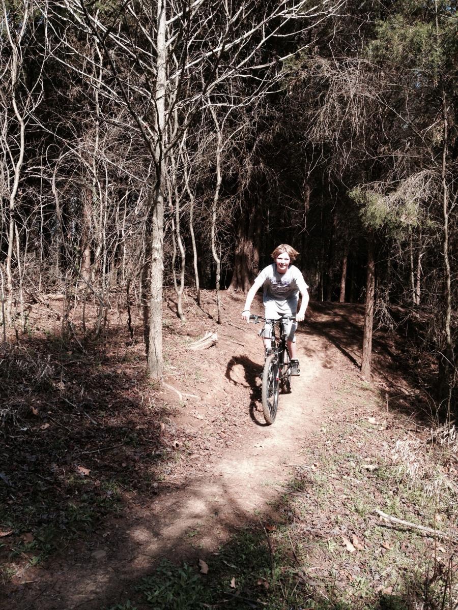 A person riding a mountain bike on a dirt trail surrounded by bare trees and foliage, smiling as they navigate a forested area in daylight. Spadra Creek Nature Trail mountain bike trail.