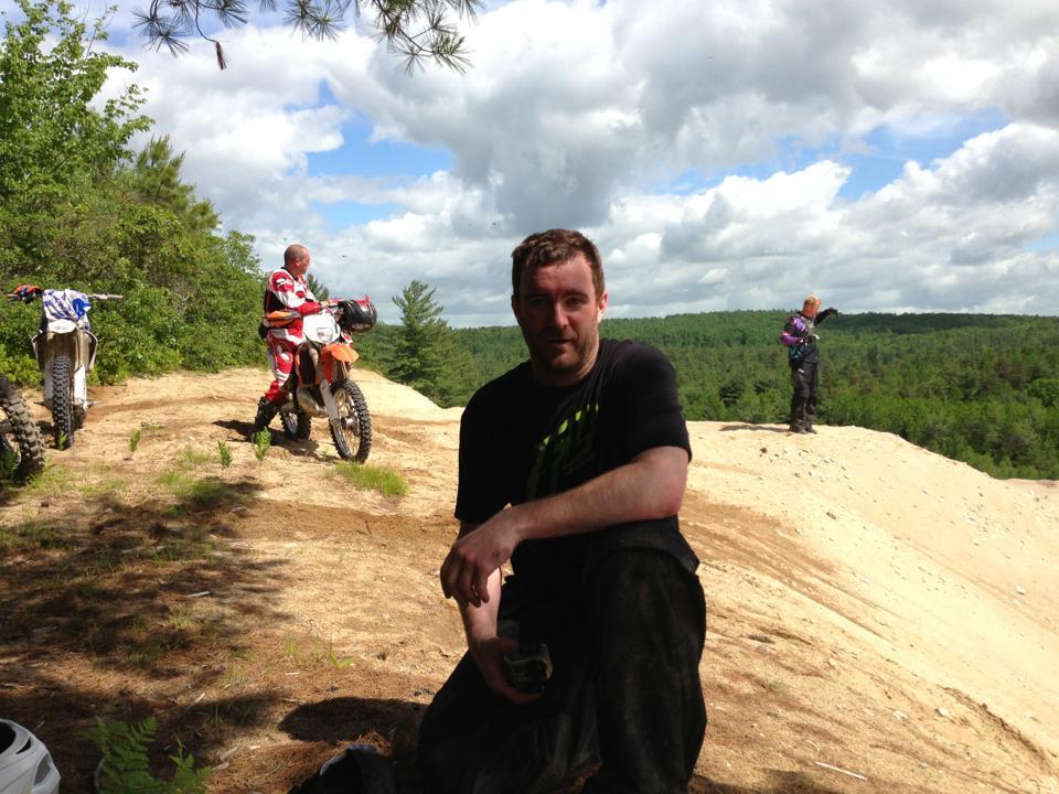 A group of three individuals enjoying a dirt biking excursion in a natural outdoor setting. One person is seated on the ground, looking at the camera, while two others stand in the background with dirt bikes parked nearby. The scene features a lush landscape with trees and a partly cloudy sky.