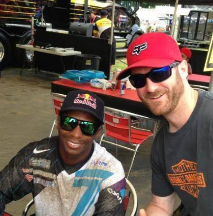 A man wearing a red hat and a dark shirt poses for a selfie with another man in a motorsport outfit and sunglasses. They are in a casual setting with tables and vehicles in the background.