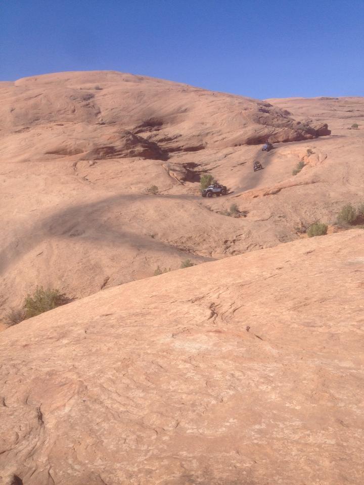 A rugged, desert landscape featuring rolling hills of reddish-brown rock. In the distance, several off-road vehicles navigate a winding trail that cuts through the rocky terrain under a clear blue sky. Sparse vegetation is visible on the rocky surface. Slickrock mountain bike trail.