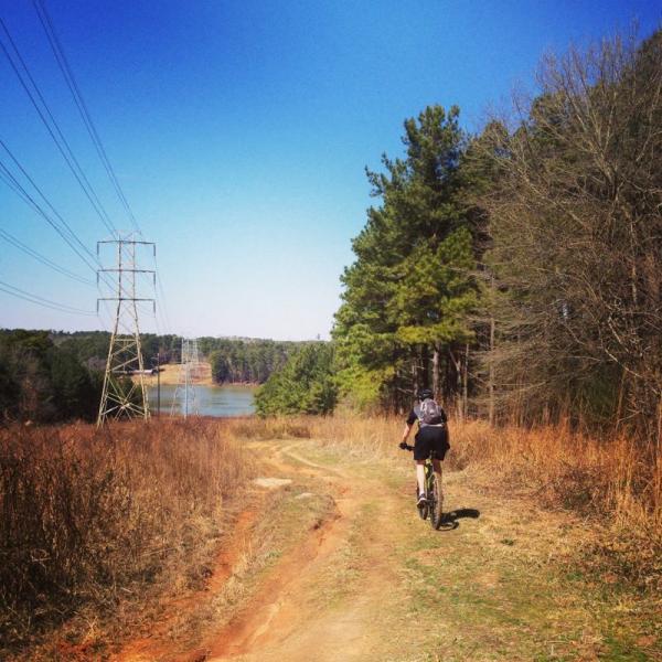 A person riding a mountain bike on a dirt trail surrounded by trees, with electrical power lines in the background and a lake visible in the distance under a clear blue sky. Fort Yargo State Park mountain bike trail.