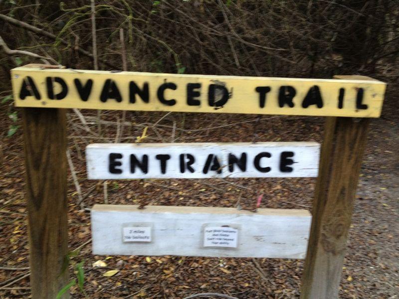 Signpost indicating the entrance to an advanced trail, with "ADVANCED TRAIL" written in black on a yellow background and "ENTRANCE" written in black on a white background. Additional information is provided on a lower wooden sign about the trail length and safety warnings. Surrounding area appears to be wooded with sparse vegetation. Caloosahatchee Regional Park mountain bike trail.