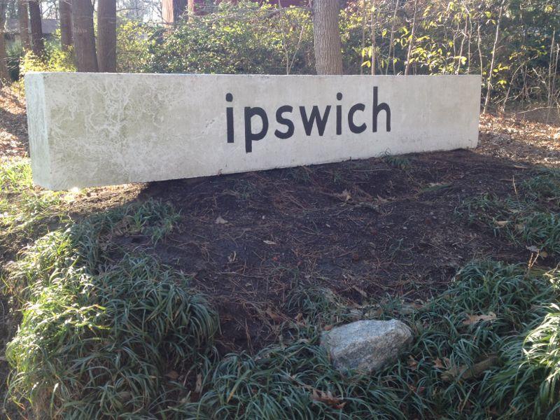 A stone sign displaying the word "ipswich" in bold black letters, surrounded by grass and small plants, set among trees and natural greenery. Indian River Park mountain bike trail.