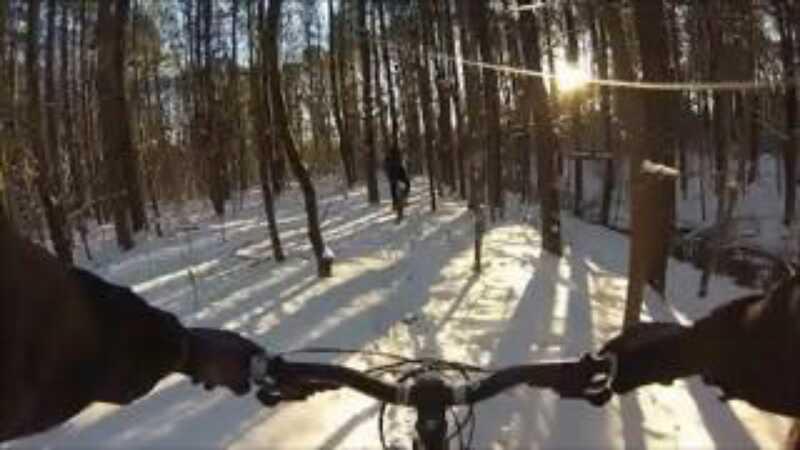 A first-person view of a mountain bike navigating through a snowy forest landscape. Sunlight filters through tall trees, casting shadows on the snow-covered ground. In the background, a person can be seen walking along a trail. Indian River Park mountain bike trail.