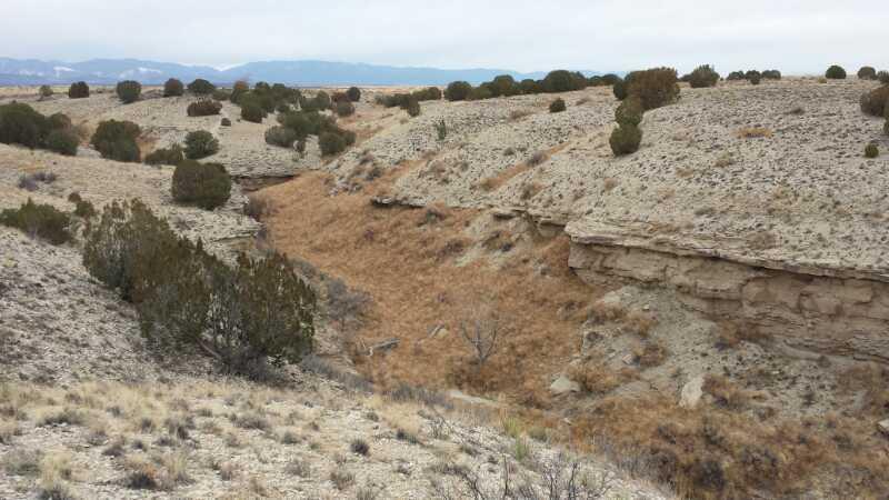 A dry, rugged landscape featuring rolling hills and a shallow valley. Sparse vegetation, including low shrubs and patches of grass, covers the ground. The scene is set against a cloudy sky, with distant mountains visible on the horizon. South Shore Lake Pueblo mountain bike trail.