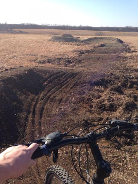 A close-up view of a mountain bike handlebar with a hand gripping it, looking towards a dirt bike path in an open field. The path has dirt mounds in the distance, indicating a trail suited for biking or jumping, with dry grass covering the surrounding area under a clear blue sky. Camp Alexander Trails mountain bike trail.