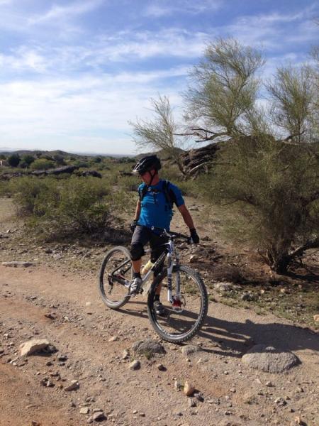 A mountain biker wearing a helmet, a blue shirt, and black shorts stands beside his bike on a rocky trail in a desert landscape. Sparse vegetation and distant hills are visible under a clear blue sky. Desert Classic mountain bike trail.
