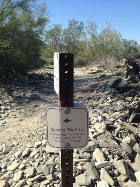 Signpost indicating the direction and distance to trails in a rocky area, labeled "Quartz Trail," with details for "Last Dog Wash Trail" and access area information. Surrounding landscape features desert vegetation under a clear blue sky. Taliesin / Quartz Loop mountain bike trail.
