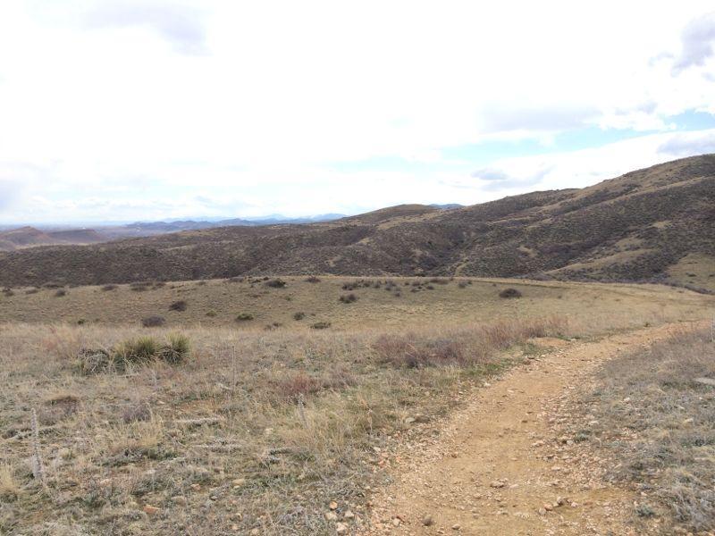 A winding dirt path leads through a grassy landscape with rolling hills, under a cloudy sky. The scene showcases a natural setting, characterized by sparse vegetation and distant mountains in the background. Devil's Backbone mountain bike trail.