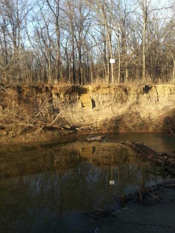 A steep, erodedbank of a river with stairs leading up to the top, surrounded by bare trees in a natural setting. The water is calm, reflecting the scenery, while a sign can be seen in the background. Pryor Creek Nature Trail mountain bike trail.