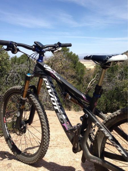 A close-up view of a mountain bike parked on a rocky terrain, with green shrubs in the background and clear blue skies above. The bike features a black frame with purple accents, front suspension forks, and knobby tires, suggesting it is designed for off-road trails. Brown Canyon mountain bike trail.