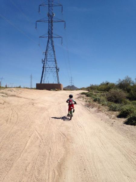 A child riding a small dirt bike on a sandy trail, with power lines and desert vegetation in the background. The clear blue sky adds to the outdoor setting. Pima Road and Dynamite Blvd mountain bike trail.