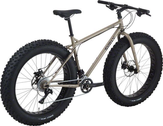 A fat tire bike with a beige frame, oversized black tires, and a single gear setup, positioned at an angle that showcases its sturdy construction and robust tires designed for off-road terrain.