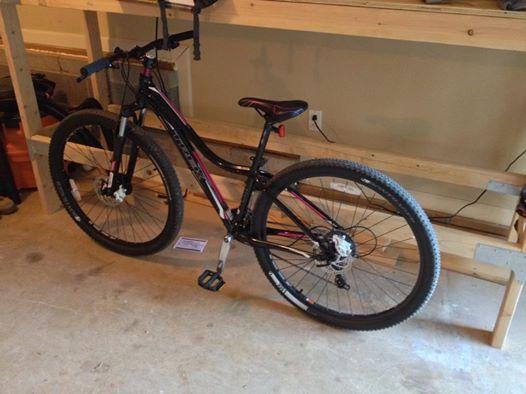 Trek Cali: A black mountain bike with pink accents is parked on a concrete garage floor. It has wide tires and is positioned next to a wooden shelving unit in the background.