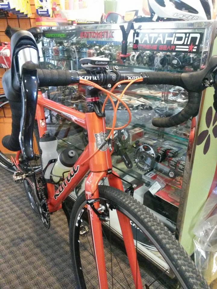 A close-up view of a bright orange bicycle displayed in a shop, featuring drop handlebars with black grips, a Garmin device mounted on the stem, and various bike components visible in the background. The shop's interior includes shelves filled with cycling accessories and equipment.