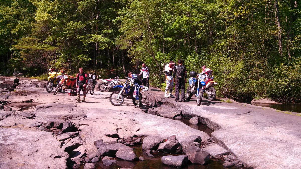 A group of off-road motorcyclists gathered on a rocky surface near a wooded area, with several dirt bikes parked nearby. The scene captures the camaraderie among the riders as they discuss their ride, surrounded by trees and nature.