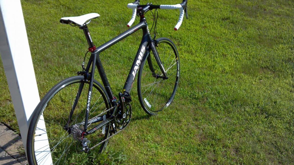 A black road bike with white handlebars, featuring a sleek frame, stands on a grassy lawn, partially leaning against a post.