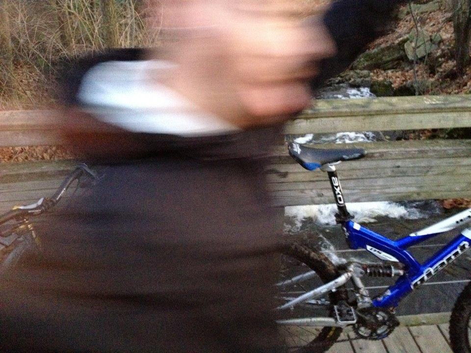 A blurry image of a person in motion next to a blue mountain bike on a wooden bridge, with a stream visible in the background. The scene captures a natural outdoor setting surrounded by trees.