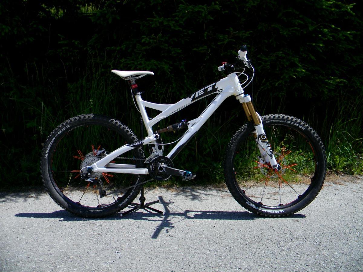 Yeti SB66: A white mountain bike with orange accents parked on a gravel path, surrounded by greenery. The bike features large, knobby tires and a sturdy frame, designed for off-road cycling.